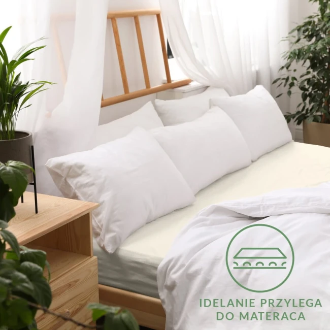 Prześcieradło jersey z gumką 120x200 w kolorze ecru leży na łóżku, złożone na materacu. Na górze widoczne poduszki w białych poszewkach.