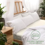Prześcieradło jersey z gumką 180x200 w kolorze ecru, leżące na łóżku z białymi poduszkami. Materiał ma delikatną teksturę. W tle drewniana rama łóżka oraz rośliny. Widoczny napis IDEALNIE PRZYLEGA DO MATERACA w dolnym rogu.