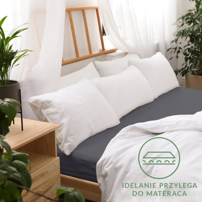 Prześcieradło jersey z gumką w kolorze ciemnym szarym o wymiarach 180x200, leżące na łóżku z białymi poduszkami. Widoczna faktura materiału, dobrze przylegająca do materaca. Tło jasne, z roślinami i drewnianymi meblami.