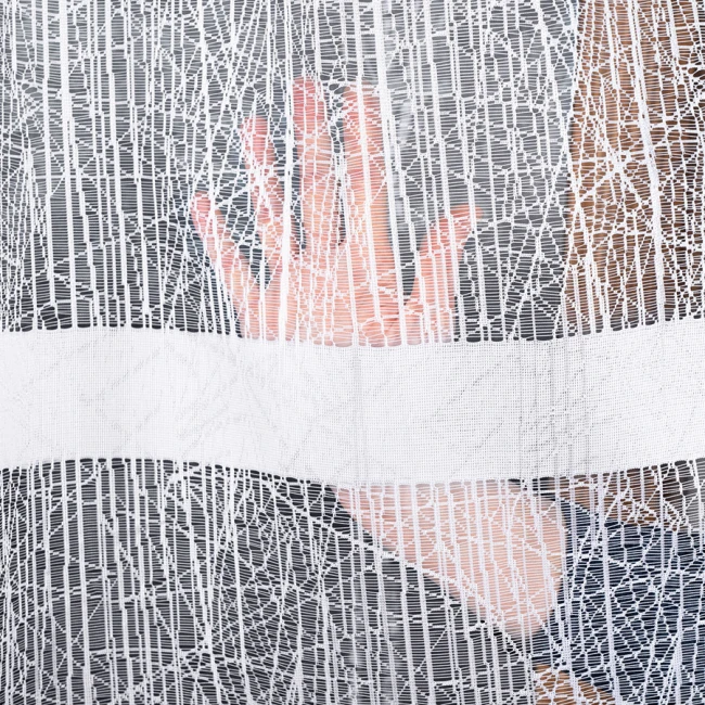 Firana żakardowa WIKTORIA w białym kolorze, rozmiar 200x250 cm, o delikatnym wzorze. Widoczna ręka z tyłu firany, kąt zbliżenia na materiał, lekko prześwitujący, wiszący na karniszu. Prosta struktura tła, wytłaczane detale.