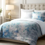 Pościel satyna bawełniana 220x200 Luxury Premium Belize w odcieniach błękitu i bieli, rozłożona na łóżku. Wzór o fakturze lejącej, z delikatnymi plamkami i nieregularnymi kształtami. W tle widoczna stolik nocny z lampą i poduszka w tym samym wzorze.