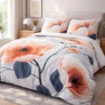 Pościel satyna bawełniana 200x200 Satynlove Premium w kolorze szarym z pomarańczowymi kwiatami, rozłożona na łóżku. Widać dwa poszewki na poduszki z dużymi motywami kwiatowymi. Tło naturalne, z zielenią roślin w pomieszczeniu.